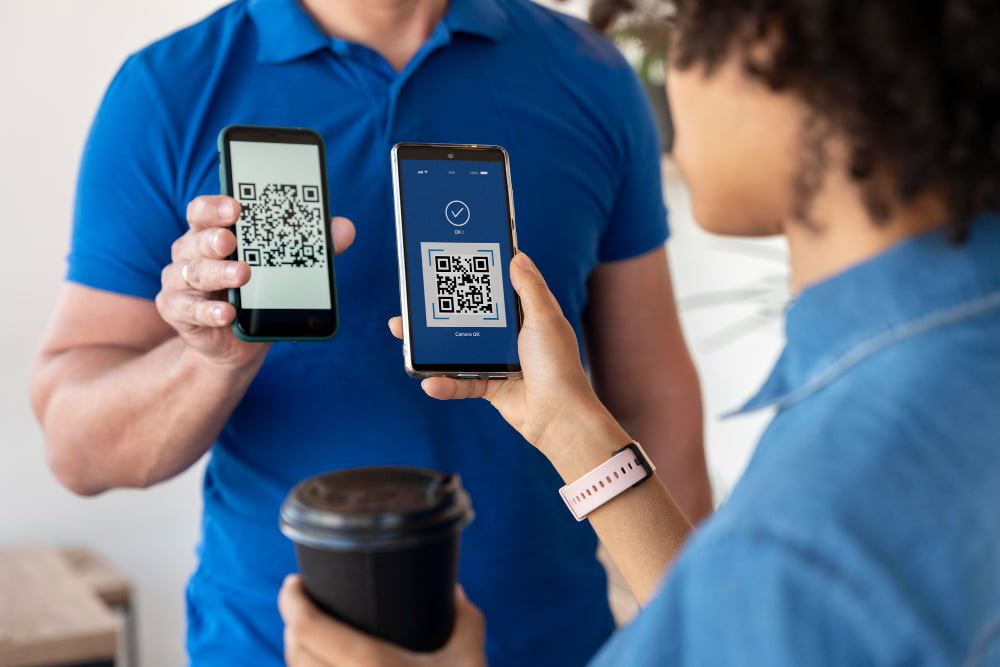 Duas pessoas estão segurando seus smartphones, cada um com um QR code na tela. Ambos parecem estar trocando informações ou verificando algo, sugerindo o uso de QR codes para compartilhamento rápido de dados.